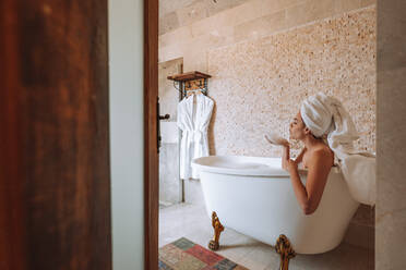 Seitenansicht einer jungen Frau mit weißem Handtuch auf dem Kopf, die sich beim Baden im hellen Badezimmer einseift - ADSF41349