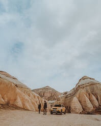Unerkanntes Paar, das in der Nähe eines gelben Geländewagens spazieren geht, der zwischen massiven Felsen gegen den bewölkten Himmel geparkt ist, während der Ferien in Kappadokien - ADSF41353