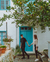 Ganzkörper-Mann und Frau in Freizeitkleidung halten sich an den Händen und schauen sich an, während sie auf einem gepflasterten Weg unter Mandarinenbaumzweigen vor einem weißen Haus mit blauer Tür und Fenstern an einem Sommerwochenende in der Türkei spazieren gehen - ADSF41358
