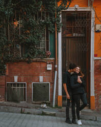 Ganzkörper-Mann, der seine Freundin umarmt und auf die Wange küsst, während er auf einem Weg in der Nähe eines orangefarbenen Hauses im Bezirk Balat in Istanbul, Türkei, steht - ADSF41362