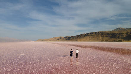 Anonymes Paar, das sich an den Händen hält, während es im rosa Wasser des Maharloo-Sees vor den Bergen und dem bewölkten blauen Himmel an einem Sommertag im Iran steht - ADSF41364