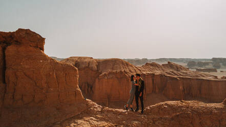Girlfriend and boyfriend in casual clothes looking at each other while standing on dry ground near ravine in desert in Iran - ADSF41366