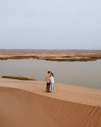 Couple standing on sandy dune and admiring calm lake on gray day in desert in Iran - ADSF41367