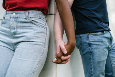 Crop unrecognizable multiracial boyfriend and girlfriend in jeans and t shirts leaning on white wall and holding hands - ADSF41368