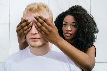 Unemotional African American girlfriend with makeup covering eyes of bearded blond boyfriend via hands with long nails while standing behind - ADSF41373