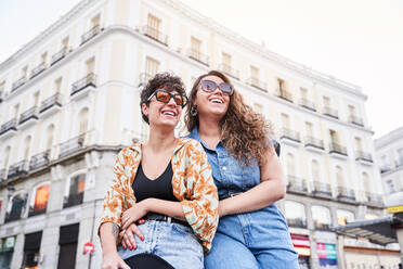 Von unten optimistische junge Freundinnen in stilvoller Kleidung und mit Sonnenbrille, die lächelnd wegschauen, während sie sich bei einem romantischen Rendezvous auf der Straße von Madrid, Spanien, umarmen - ADSF41376