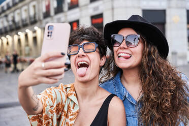 Happy young girlfriends showing tongues and taking selfie via smartphone on weekend day on street on Madrid, Spain - ADSF41378