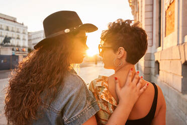 Back view woman in hat and sunglasses touching girlfriend during romantic date at sunset on street of Madrid, Spain - ADSF41381