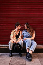 Full body stylish young girlfriends smiling and looking at each other while sitting on border near red wall on street of Madrid, Spain - ADSF41392