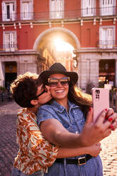 Young woman hugging and kissing happy girlfriend while taking selfie on sunlit street at sunset in Madrid, Spain - ADSF41398