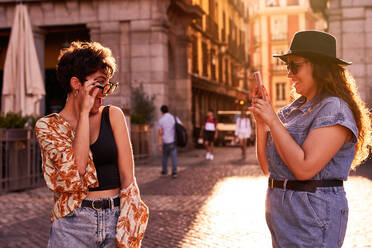 Glückliche junge Frau in Freizeitkleidung mit Hut und Sonnenbrille, die lächelt und ihre stilvolle Freundin bei einem romantischen Rendezvous bei Sonnenuntergang auf einer Straße in Madrid, Spanien, fotografiert - ADSF41400