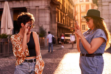 Happy young female in casual clothes with hat and sunglasses smiling and taking photo of stylish girlfriend during romantic date at sunset on street of Madrid, Spain - ADSF41400