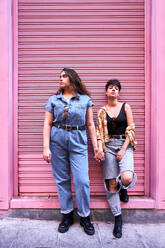 Ganzkörper stilvolle junge Freundinnen halten sich an den Händen und lehnen an der rosa Wand, während sie Zeit auf der Straße von Madrid, Spanien verbringen - ADSF41405