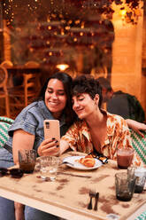 Happy young girlfriend smiling and browsing social media on cellphone while sitting at table during date in street cafeteria in Madrid, Spain - ADSF41410