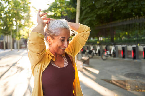Positiv eingestellte ältere Sportlerin, die lächelt und sich ein Haargummi um die grauen Haare bindet, während sie sich auf ein Fitnesstraining auf einer sonnenbeschienenen Straße in der Stadt am Morgen vorbereitet - ADSF41417