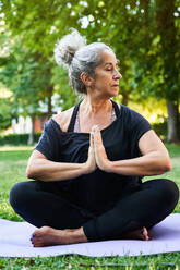 Ruhige ältere Frau in Sportkleidung sitzt mit geschlossenen Augen auf einer Yogamatte und übt Padmasana, während sie an einem sonnigen Tag im Park meditiert - ADSF41419