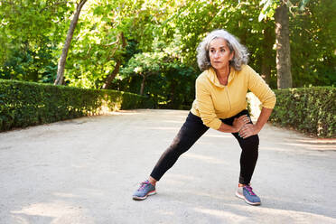 Elderly female athlete with gray hair stretching legs in park - ADSF41423
