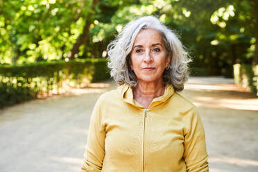 Happy aged female with gray hair looking away with smile while standing near hedge on sunny summer day in park - ADSF41425