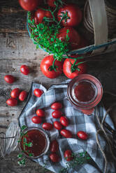 Draufsicht auf reife Tomaten und grüne Kräuter in einem Korb neben einer karierten Serviette und Gläsern mit Marmelade auf einem Küchentisch aus Holz - ADSF41449