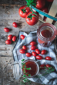 Draufsicht auf reife Tomaten und grüne Kräuter in einem Korb neben einer karierten Serviette und Gläsern mit Marmelade auf einem Küchentisch aus Holz - ADSF41450