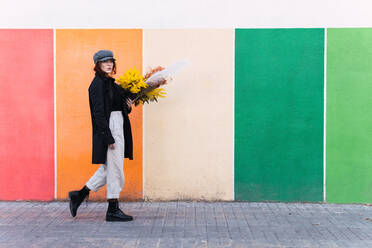 Ganzkörper stilvolle junge Frau in Oberbekleidung trägt Strauß frischer Blumen und Blick auf die Kamera beim Schlendern auf dem Bürgersteig gegen bunte Wand in der Stadt Straße - ADSF41454
