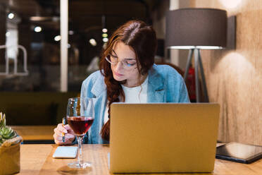 Intelligente junge Frau mit Brille, die Daten auf einem Smartphone liest und analysiert, während sie am Tisch mit Gadgets und Rotwein während der Arbeit in einem Restaurant am Abend sitzt - ADSF41455