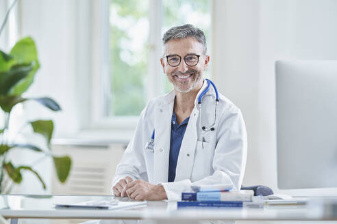 Happy mature doctor in lab coat sitting in medical practice - RORF03075