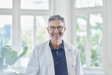 Happy mature doctor wearing eyeglasses standing in medical practice - RORF03108