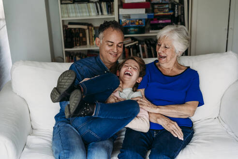 Happy girl with father and grandmother enjoying on sofa at home - EBBF07255