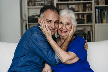 Smiling son and mother sitting with cheek to cheek on sofa at home - EBBF07260