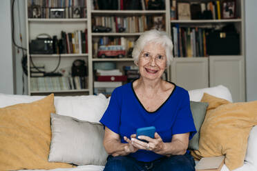 Glückliche ältere Frau mit Smartphone auf dem Sofa zu Hause - EBBF07262