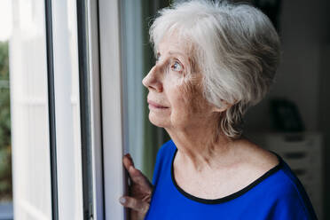 Thoughtful senior woman looking through window at home - EBBF07264