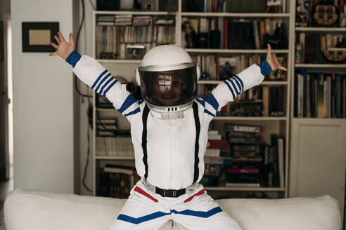 Happy girl wearing space costume sitting with arms outstretched on sofa - EBBF07268