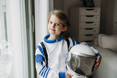 Mädchen im Weltraumkostüm mit Helm steht am Fenster zu Hause - EBBF07280