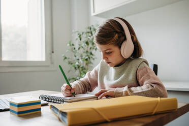 Mädchen mit drahtlosen Kopfhörern macht Hausaufgaben am Tisch sitzend zu Hause - EBBF07310