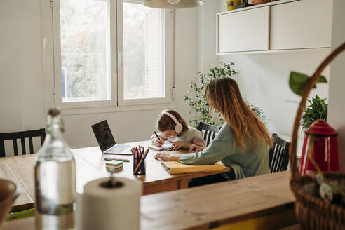 Mutter sieht ihre Tochter bei den Hausaufgaben an, die mit einem Laptop am Tisch sitzt - EBBF07321