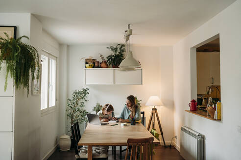 Mother explaining daughter sitting with laptop at table at home - EBBF07322