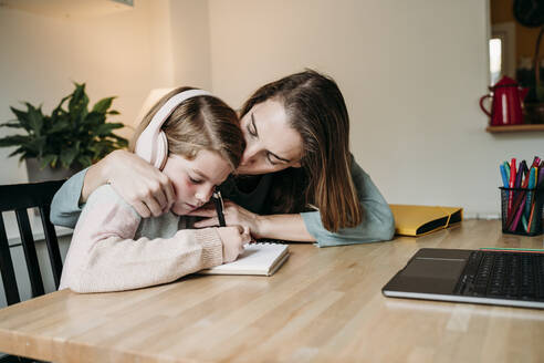 Mother kissing daughter doing homework at table at home - EBBF07325