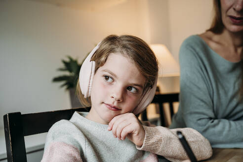 Cute thoughtful girl on chair sitting by mother at home - EBBF07326