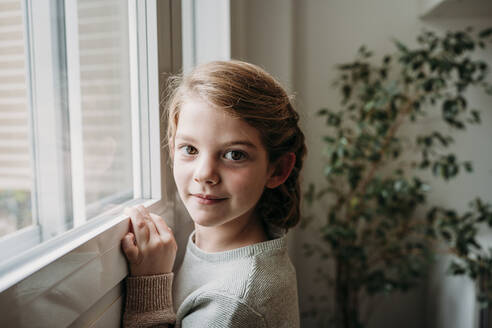 Smiling cute girl standing by window at home - EBBF07330