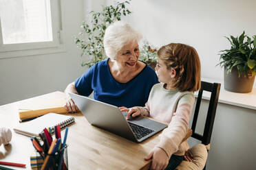Glückliches Mädchen mit Laptop auf dem Tisch und Blick auf die Großmutter - EBBF07334