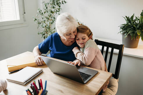 Smiling girl with laptop on table embracing grandmother at home - EBBF07335