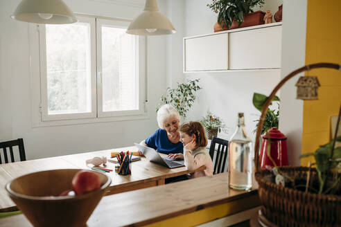 Happy senior woman helping granddaughter studying through laptop on table at home - EBBF07338
