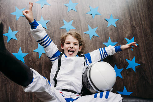 Playful girl wearing space costume lying by helmet and blue stars on floor at home - EBBF07348