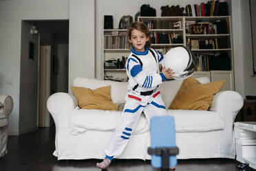 Girl wearing space costume holding helmet dancing and recording video on mobile phone at home - EBBF07363