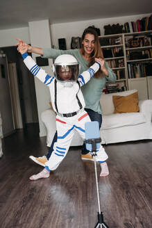Daughter wearing space costume dancing with mother and recording on mobile phone at home - EBBF07368