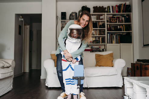 Mother with daughter wearing space costume taking selfie through mobile phone at home - EBBF07369