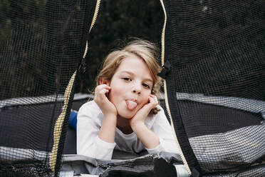 Mädchen streckt die Zunge heraus und liegt auf einem Trampolin im Garten - EBBF07372