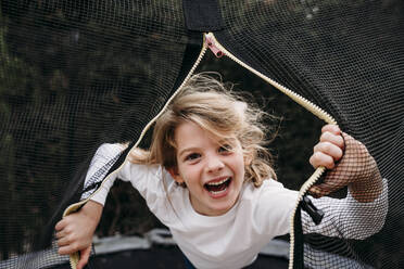 Glückliches Mädchen schaut durch das Netz auf dem Trampolin im Garten - EBBF07374