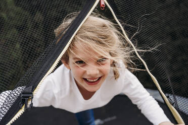 Glückliches süßes Mädchen schaut durch Netz auf Trampolin im Garten - EBBF07375